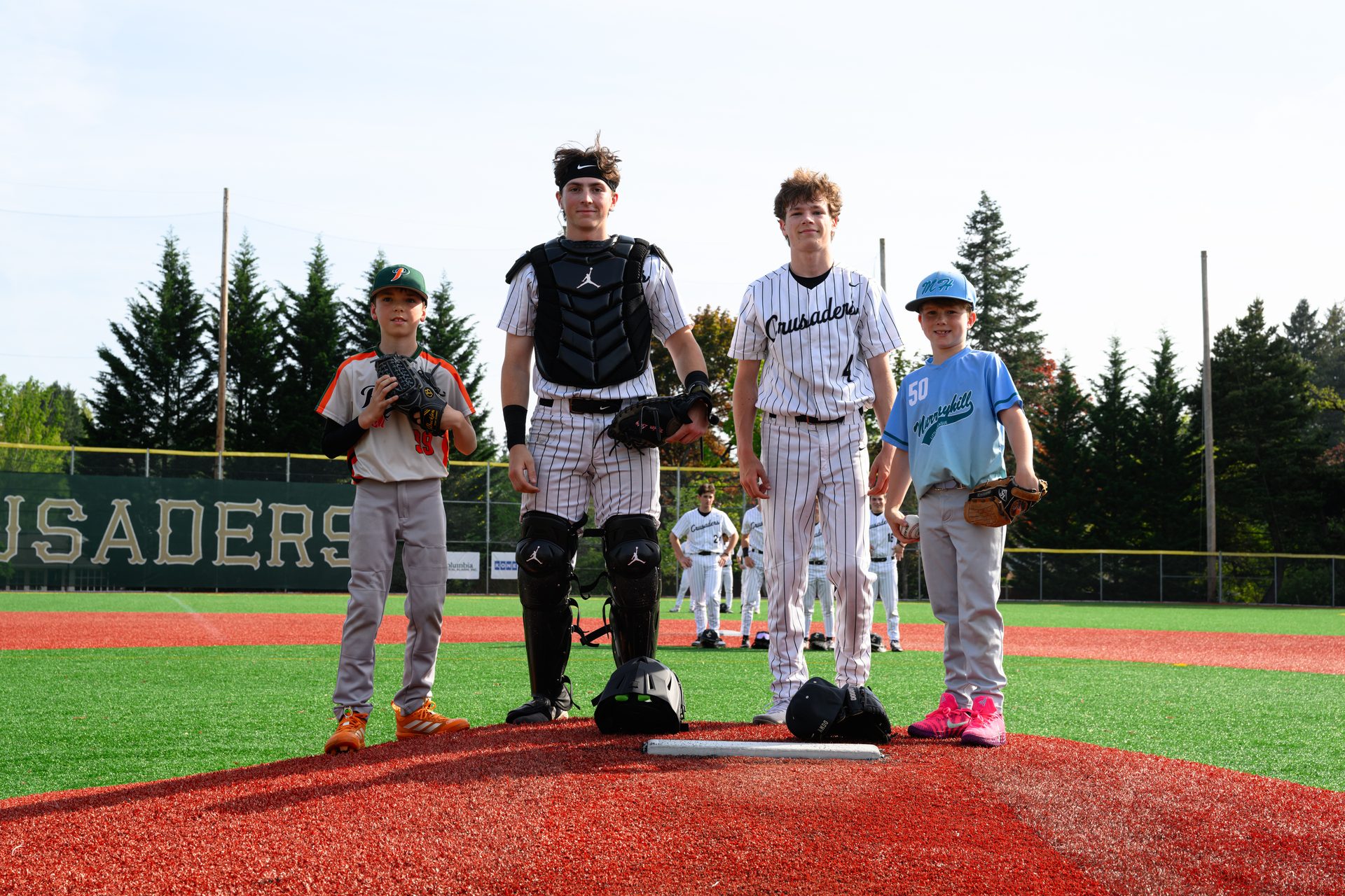 Four young players of different ages stand together on the mound, gloves in hand, ready for the game to begin. Jesuit High School Baseball, 2026.