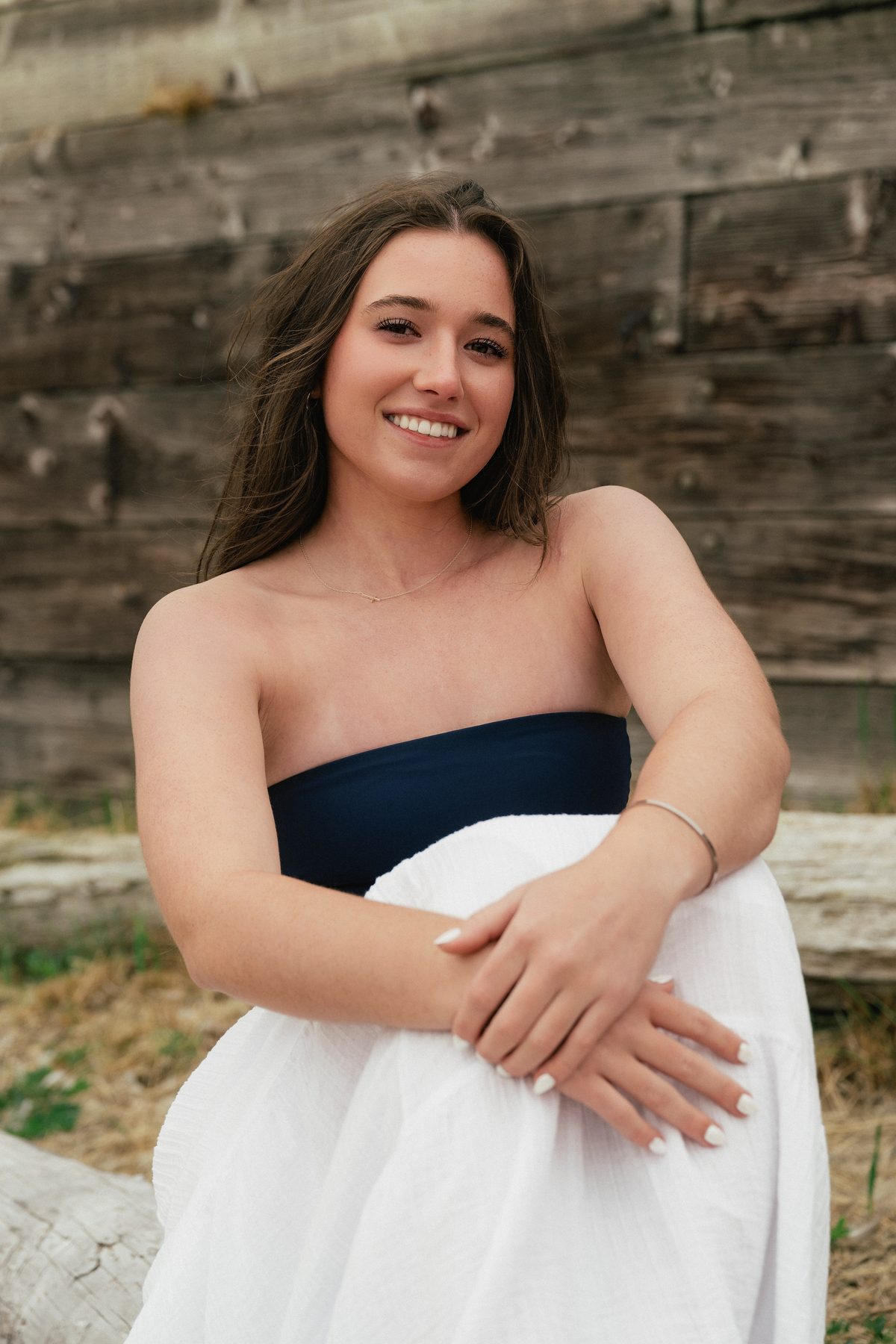 She sits against the rustic wood wall, arms folded gently over her knee, sharing an easy, genuine smile. Jesuit High School Senior Portrait, 2026.