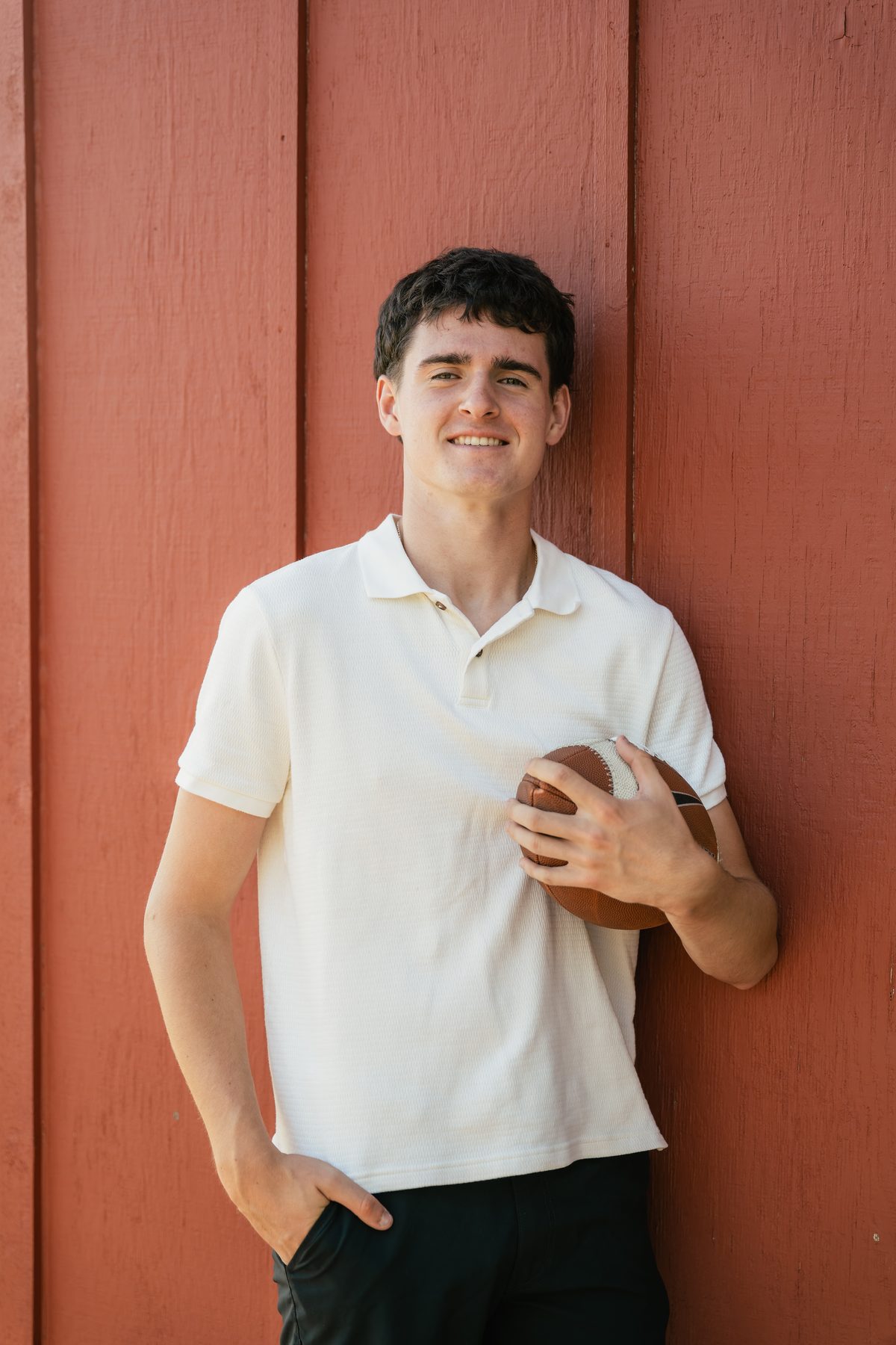 He grins at the camera, football tucked casually under one arm against a red barn wall. Jesuit High School Senior Portrait, 2026.