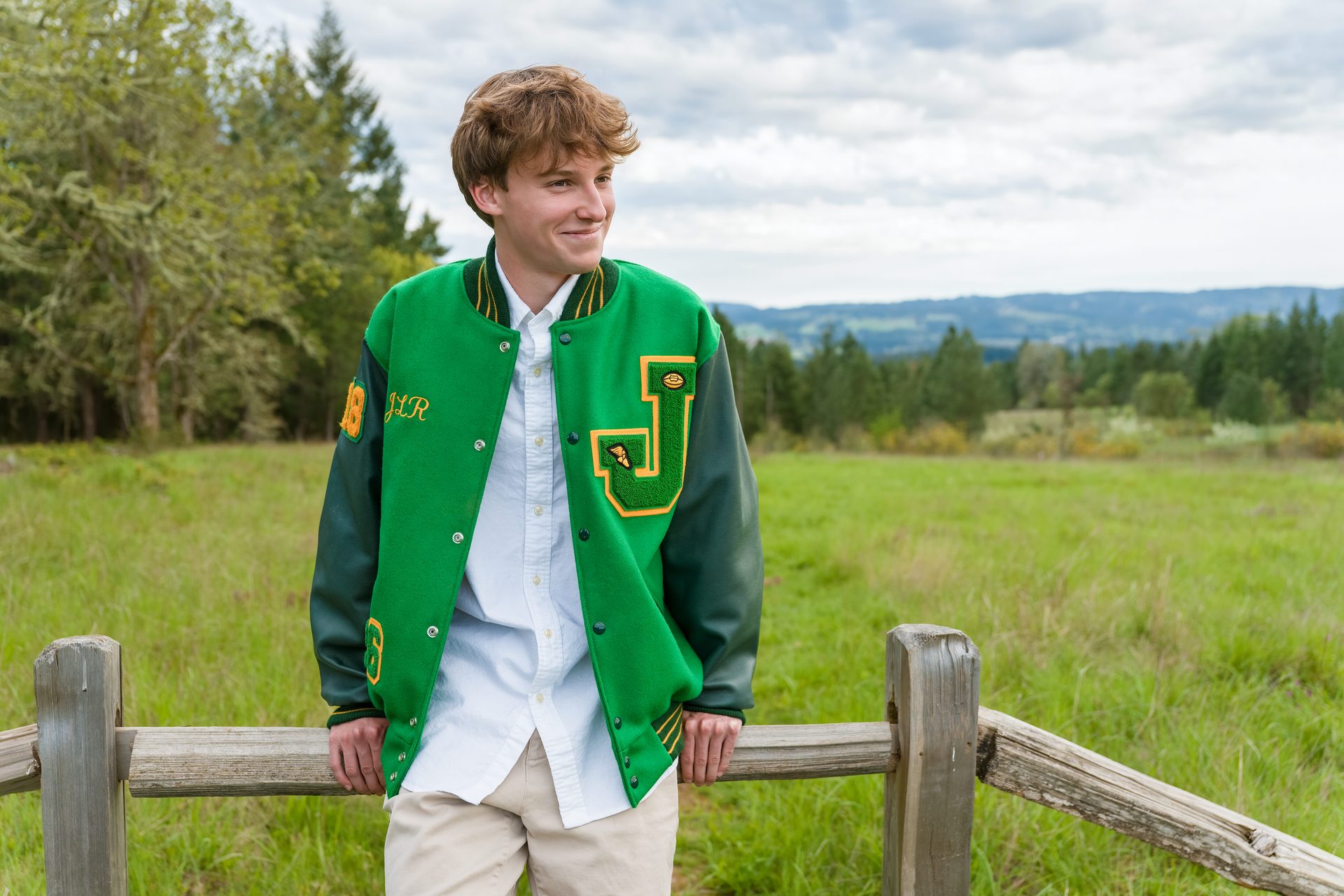 He leans against the old fence rail, smiling off to the side while the wide green field stretches out behind him under a soft gray sky. High School Senior Portrait, 2026.