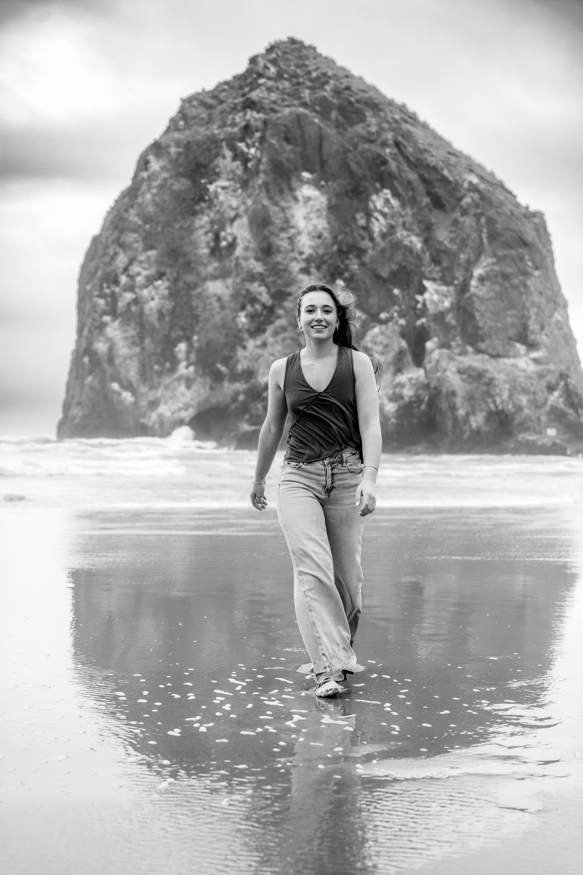 She walks barefoot across the wet sand, smiling, with a giant rock formation and rolling waves filling the scene behind her. Jesuit High School Senior Portrait, 2026.