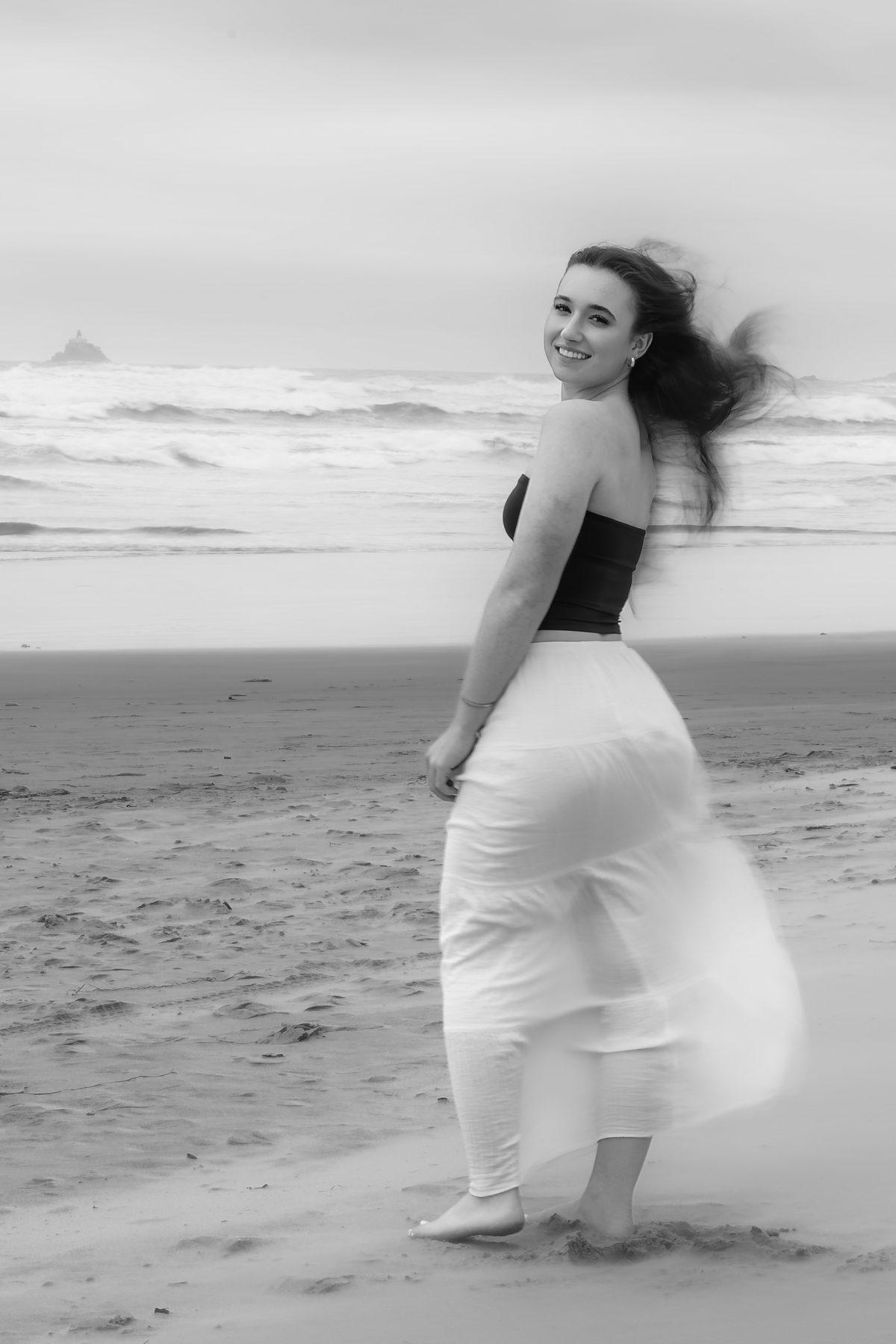 Her hair and skirt catch the wind as she turns back with a smile, the waves rolling in softly behind her. Jesuit High School Senior Portrait, 2026.