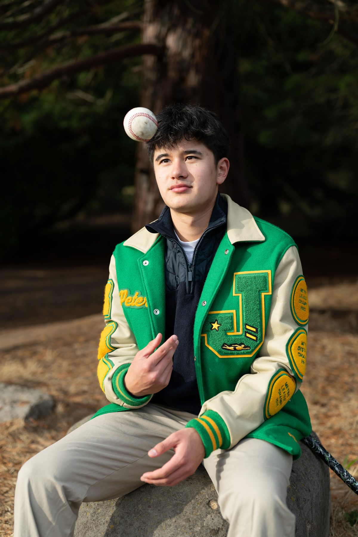 A young athlete in a varsity letterman jacket sits on a rock outdoors, watching a baseball hover in the air above him. Jesuit High School Senior Portrait, 2026.