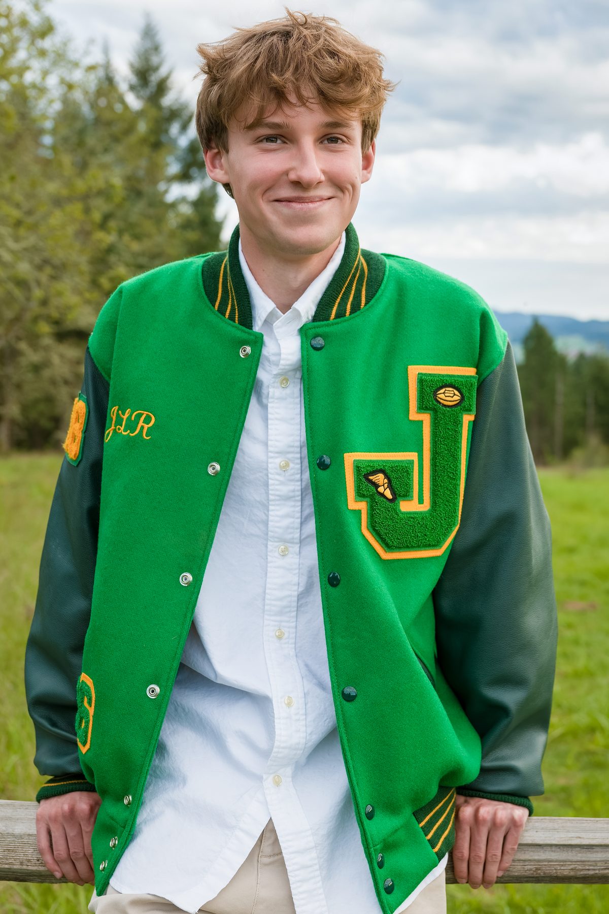 He rests his arms on a wooden rail, wearing his letterman jacket with a relaxed, easy smile. Jesuit High School Senior Portrait, 2026.