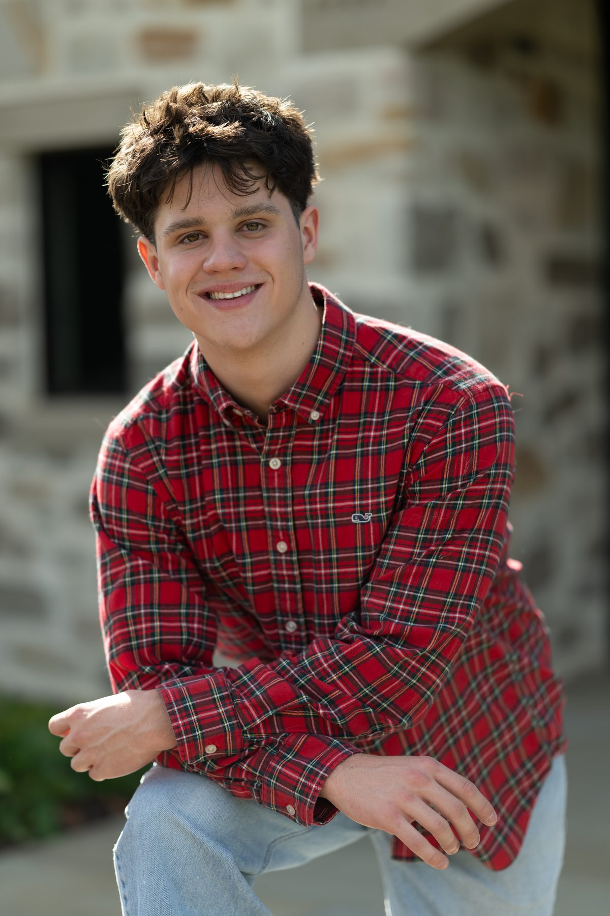 He leans forward with a relaxed grin, his red plaid shirt bright against the muted stone wall behind him. Jesuit High School Senior Portrait, 2026.