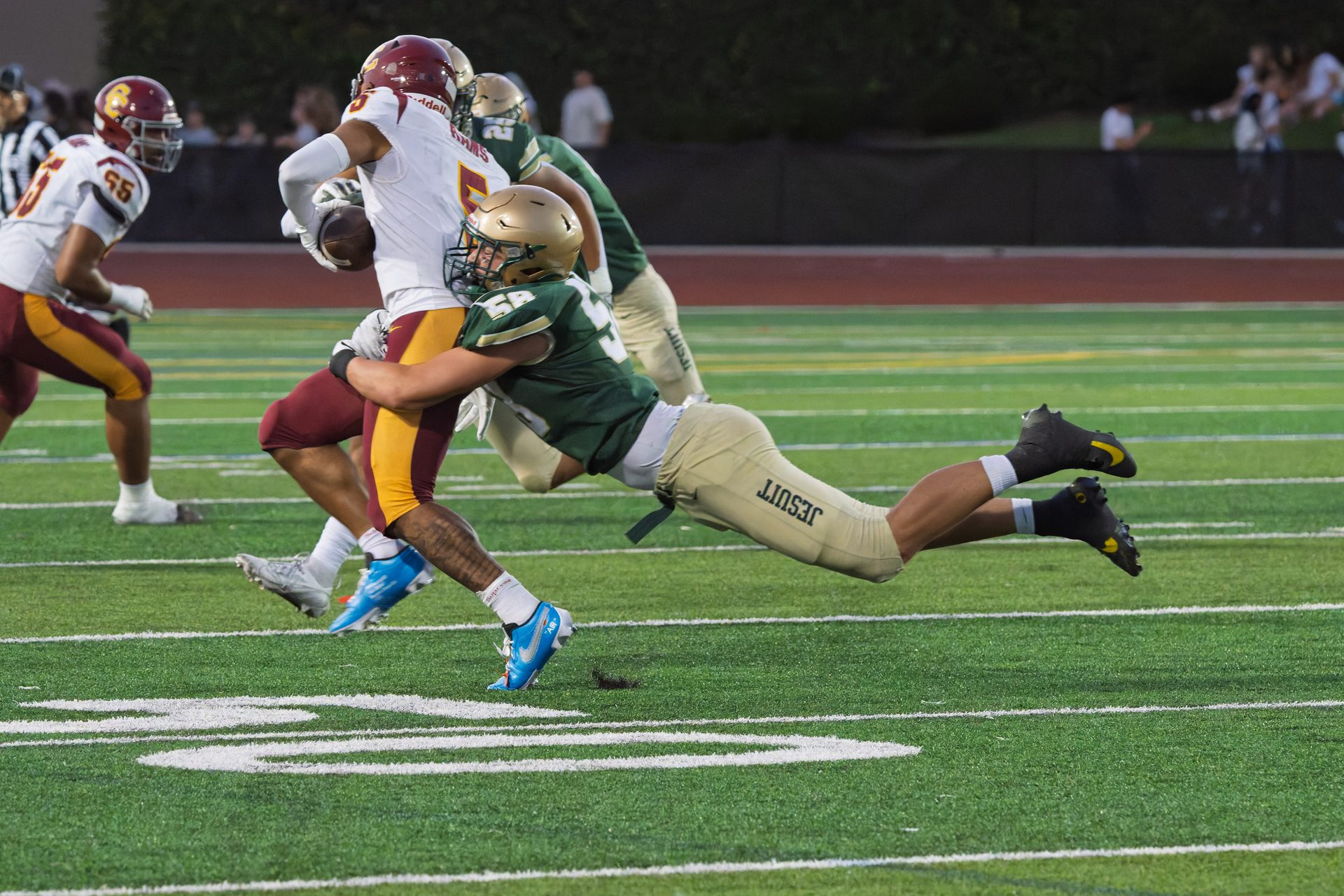A defender launches fully off the ground to wrap up the runner near midfield as teammates close in from behind. Jesuit High School Football, 2026.