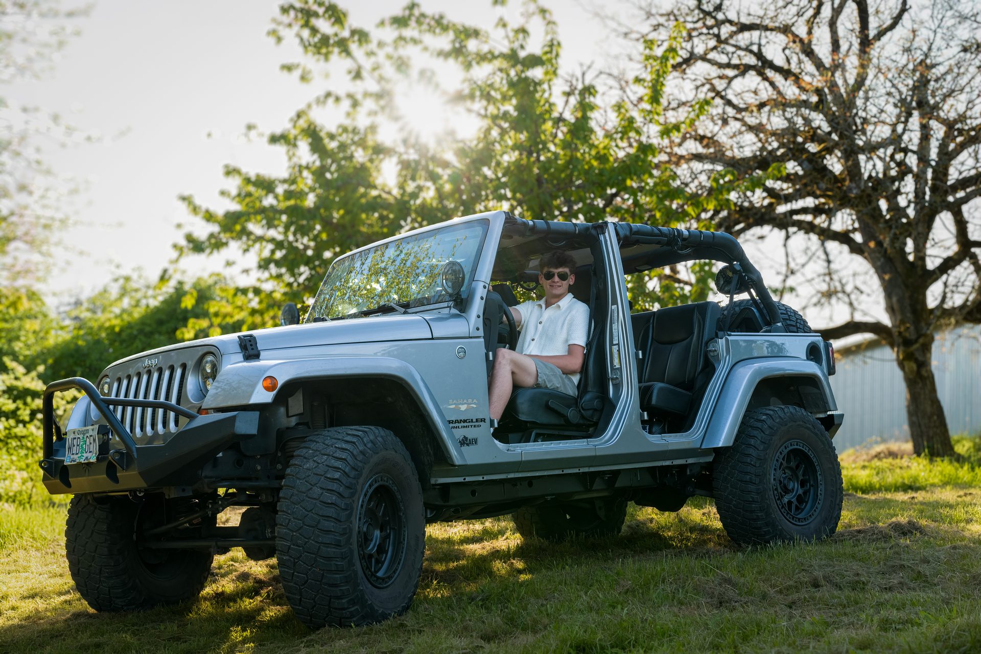 A young man in sunglasses relaxes behind the wheel of an open-air off-roader as sunlight filters through the trees behind him. High School Senior Portrait, 2026.